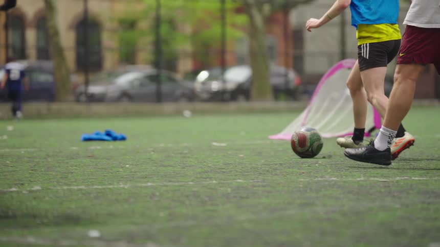 A young man kicks a soccer ball in slow motion during a competitive pickup game with friends in a city park