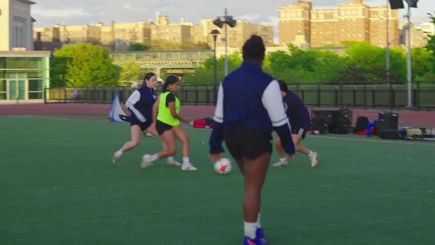 A diverse group of young women practice their soccer skills in slow motion on a turf field in an urban setting