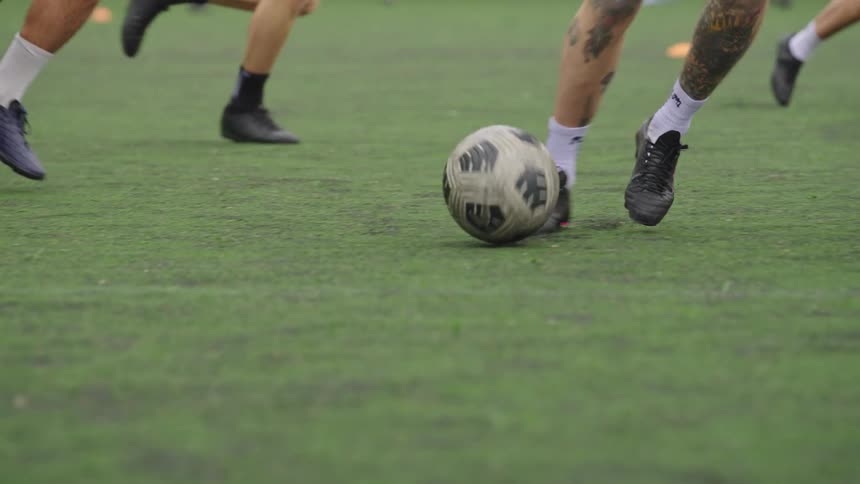 A dynamic slow-motion close-up follows a soccer ball as players' feet chase it across an artificial turf field