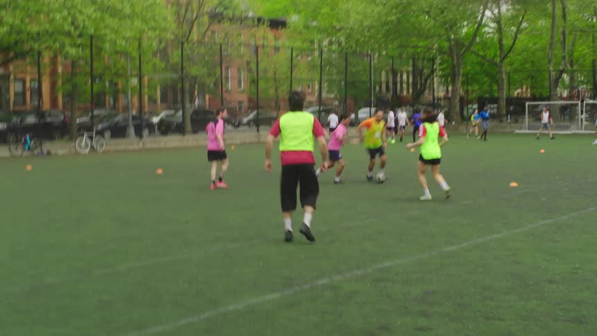 A diverse group of friends plays a casual soccer game on a green turf field in a city park in slow motion