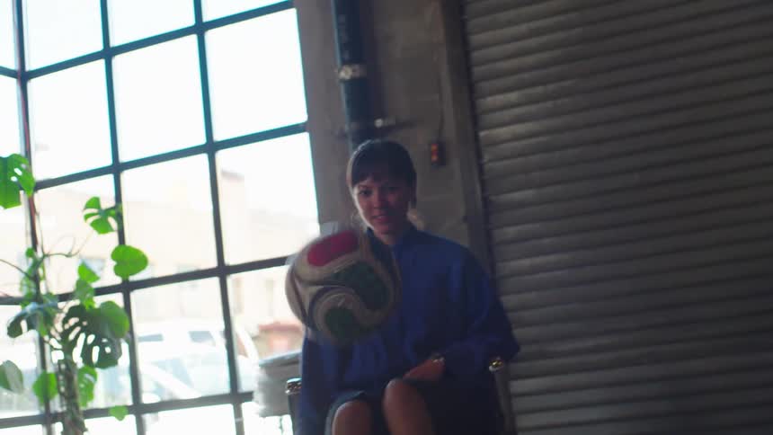 A woman in a creative loft office juggles a soccer ball with her feet while sitting in a chair, then gets up and walks to the window