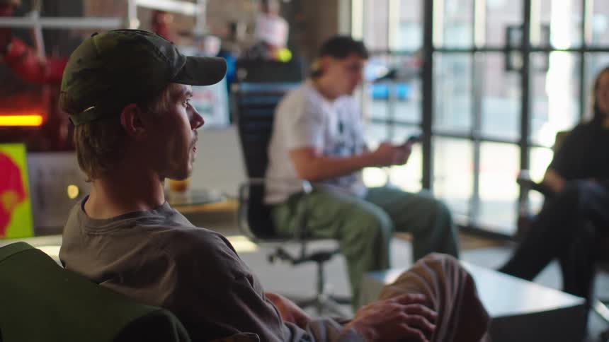 A group of young adults hang out in a sunlit industrial loft, with the camera pulling focus between two men on their phones