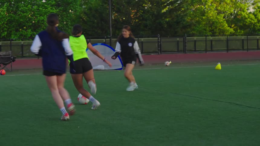 In slow motion, a young female soccer player kicks and dribbles the ball down the field during a team practice at dusk