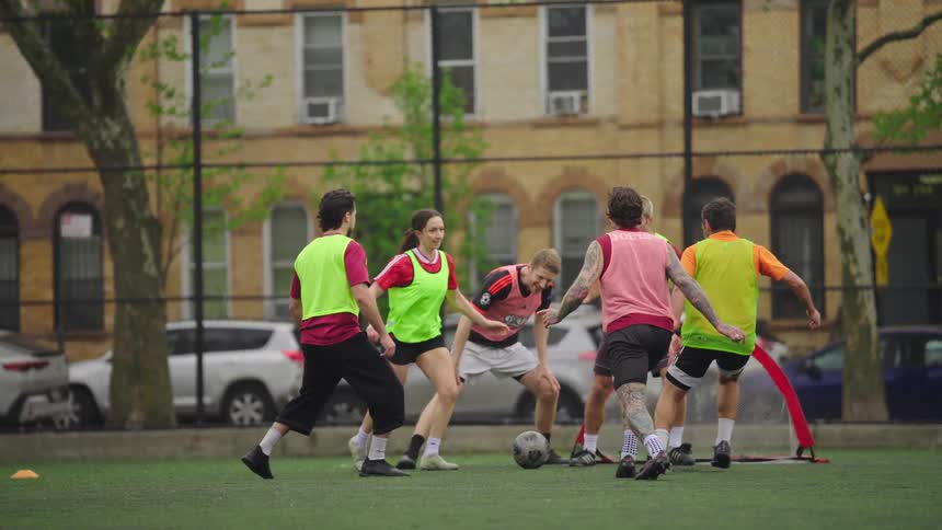A group of athletic friends play a competitive game of co-ed soccer on an urban field in slow motion