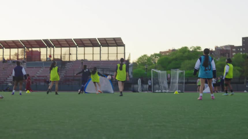 Slow motion shot of a girls soccer team practicing drills on an urban field with city buildings in the background at sunset