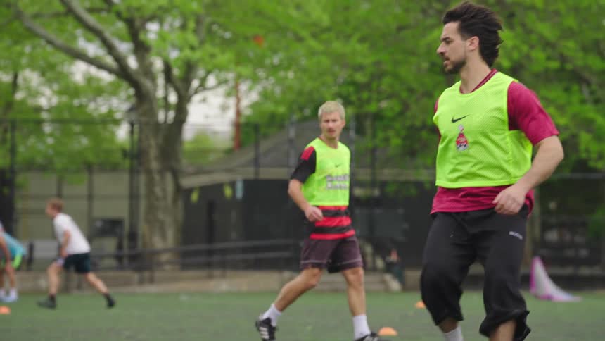 A man in a yellow vest dribbles a soccer ball past a defender in slow motion during a pickup game in a city park
