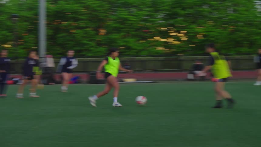 A young woman in a neon vest dribbles a soccer ball during a team practice on a city field at dusk
