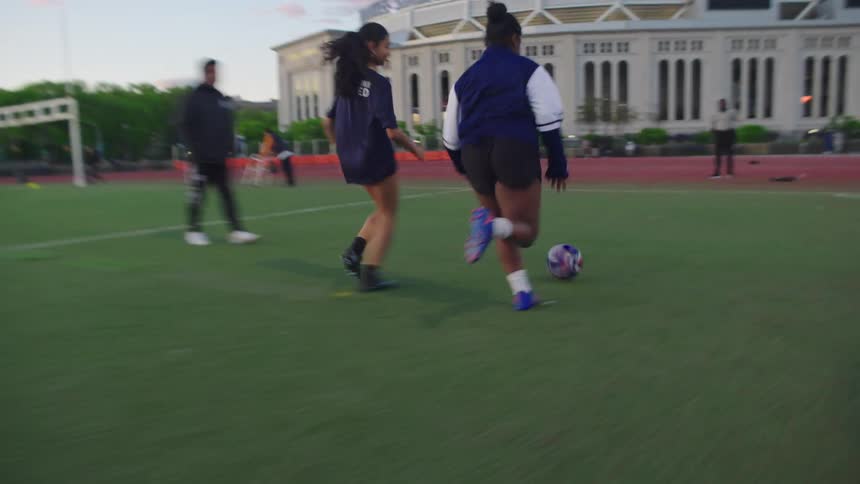 A young female soccer player skillfully dribbles past a defender in slow motion during a practice session on a city field with Yankee Stadium in the background