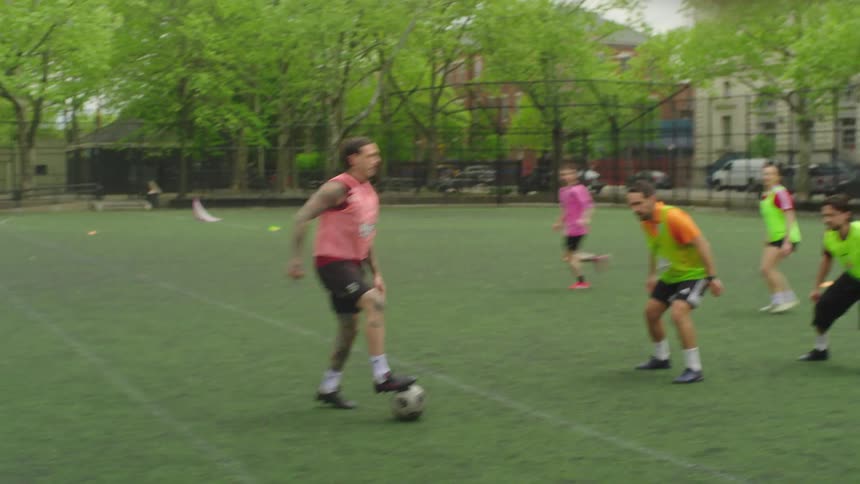 Dynamic slow-motion footage follows players as they dribble and pass during a fast-paced soccer game in a city park