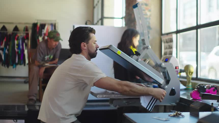 A man in a design studio uses a heat press machine to print a custom design onto a t-shirt in slow motion