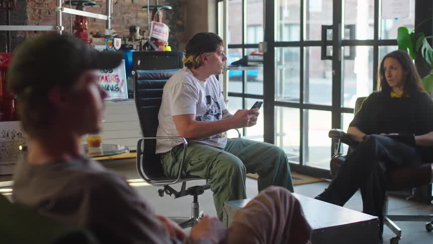 A young man talks animatedly while the focus shifts to his two friends hanging out in a modern industrial loft office