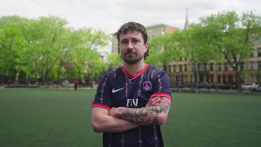 A confident tattooed man in a soccer jersey stands with his arms crossed on an urban park field as the camera slowly zooms in for a portrait
