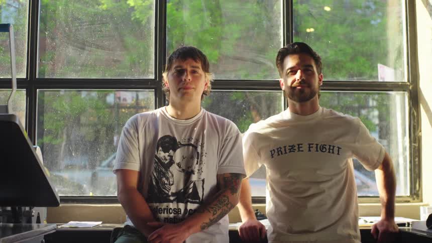 A slow push-in portrait of two confident male business partners standing in front of a large window in their sunlit workshop