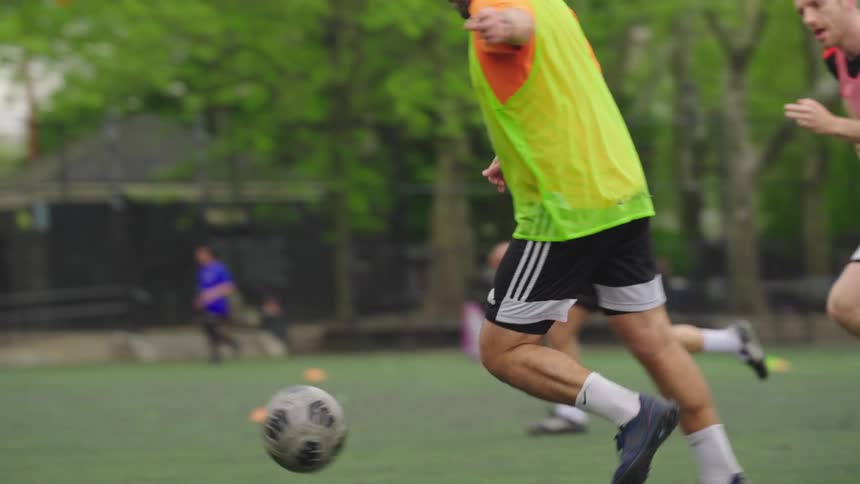 A soccer player's feet kick a ball in slow motion during a pickup game on an urban turf field