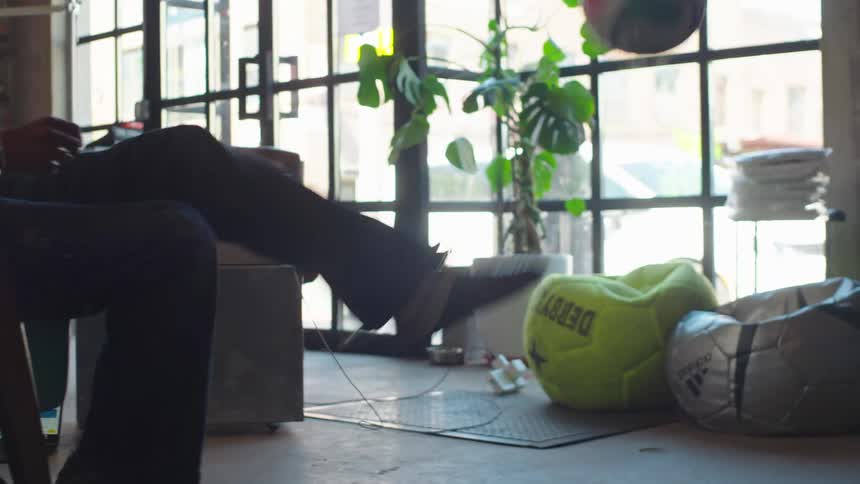 A man and a woman skillfully juggle a soccer ball with their feet while seated in a sunlit industrial loft