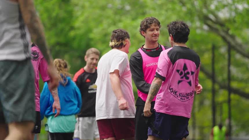 A group of soccer players wearing pink jerseys talk and laugh together while standing on an outdoor field