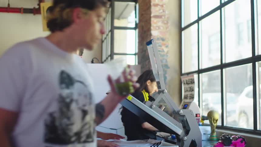 A man carefully prepares a jersey on a heat press machine in a sunlit clothing design workshop, smiling as a coworker walks by