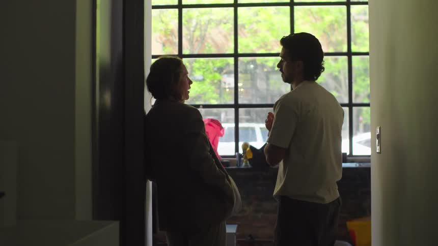 A silhouetted man and woman engage in a quiet conversation in a dark hallway, framed by a large sunlit window