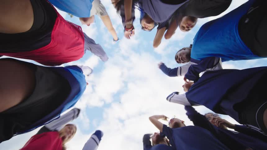 Low angle view of a diverse sports team huddling in a circle, putting their hands together and cheering in unison under a cloudy sky