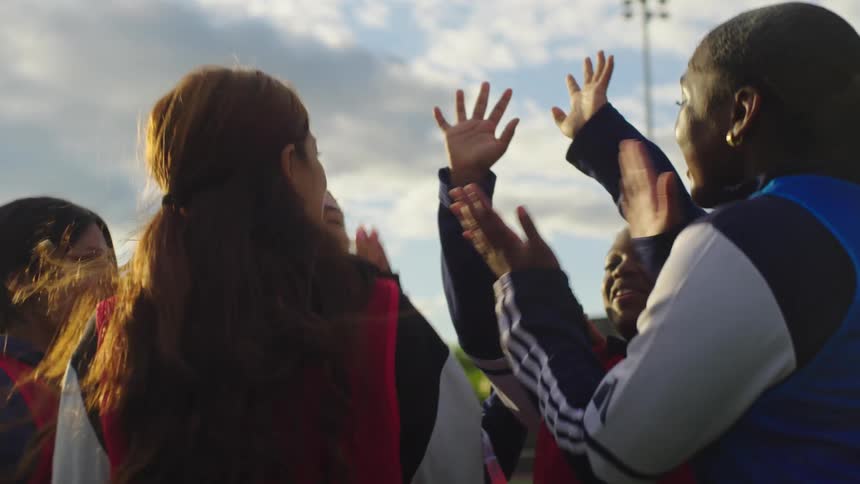 An energetic and diverse team of young women athletes huddle together, hugging and cheering with their hands in the air on a sunny sports field
