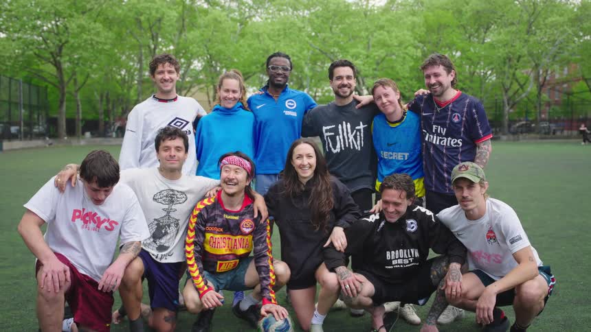 A diverse co-ed soccer team smiles and poses for a group photo on a turf field in a city park