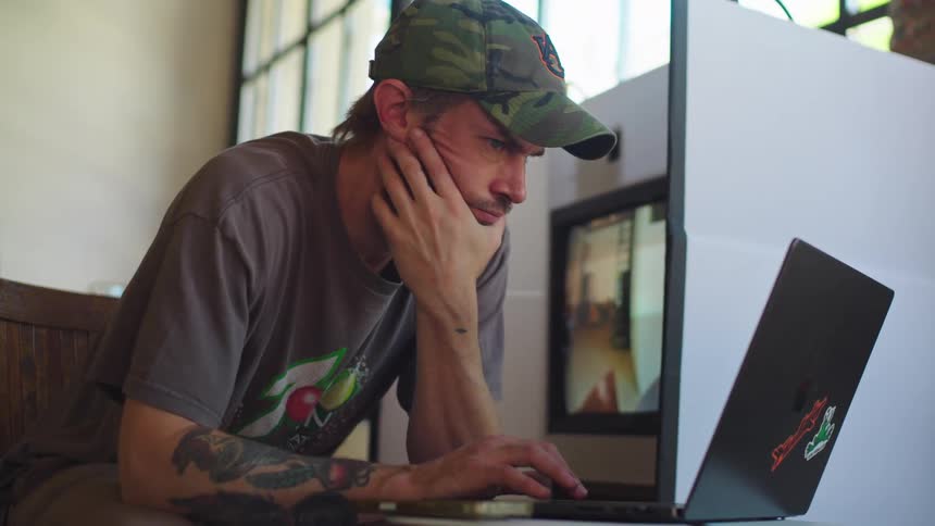 A tattooed young man in a camouflage baseball cap sits intently focused on his laptop screen while working in a sunlit room