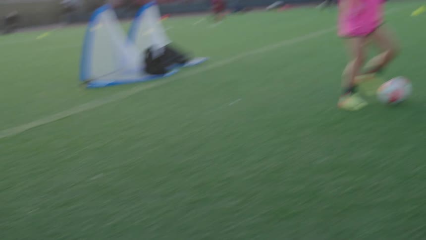 A slow-motion tracking shot follows a young female soccer player dribbling the ball during team practice on a city field