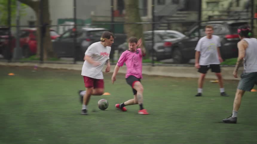 A group of tattooed young men play a casual game of soccer in an urban park, captured in slow motion