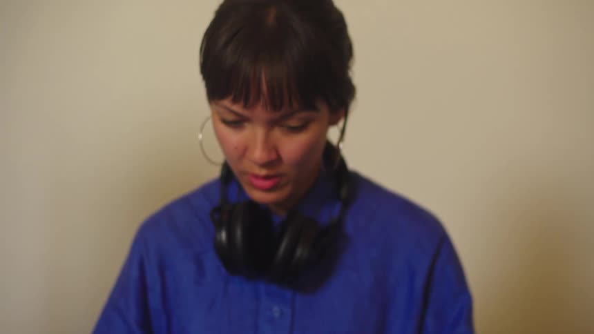 A female DJ with headphones concentrates on her mixing console, with the camera starting on her hands and tilting up to her face