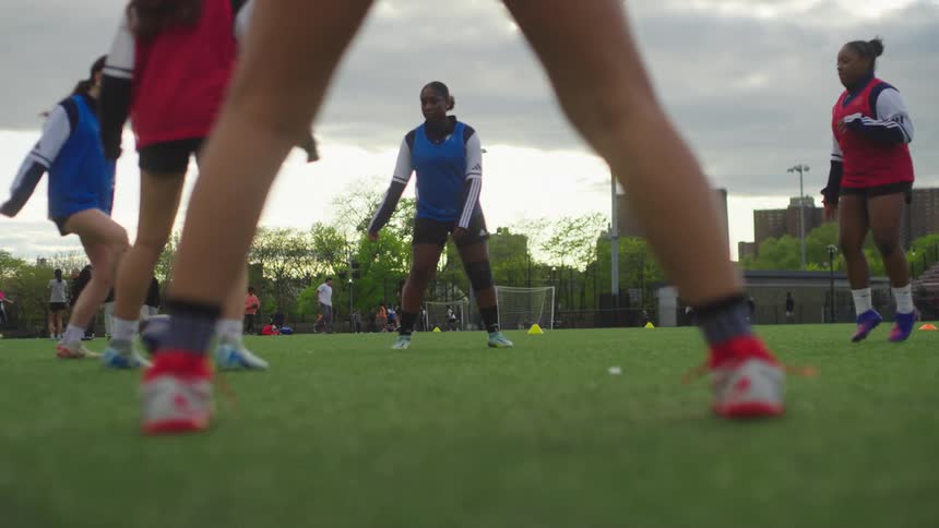A slow-motion, low-angle view shows female soccer players' feet as they practice passing drills on an urban turf field