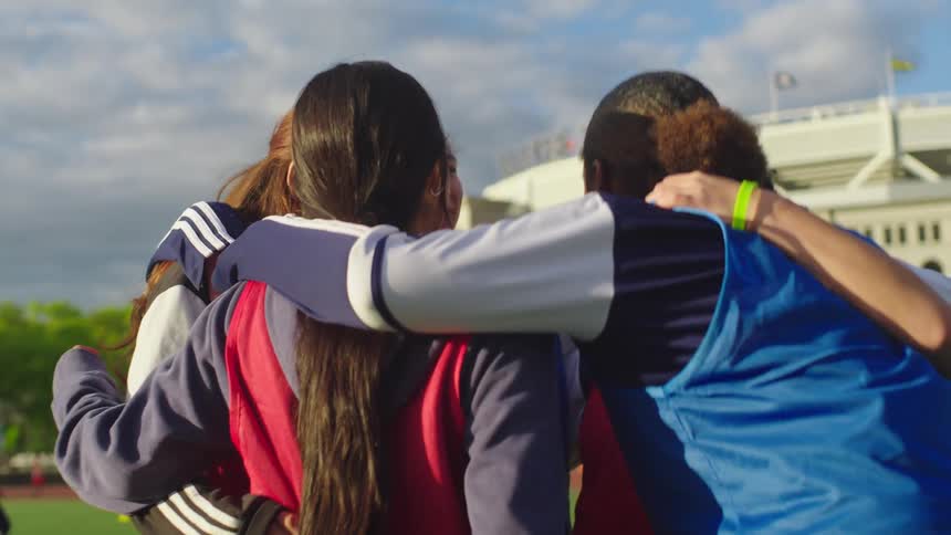 A diverse girls' soccer team shares a moment of unity, smiling and coming together for a team huddle in front of Yankee Stadium