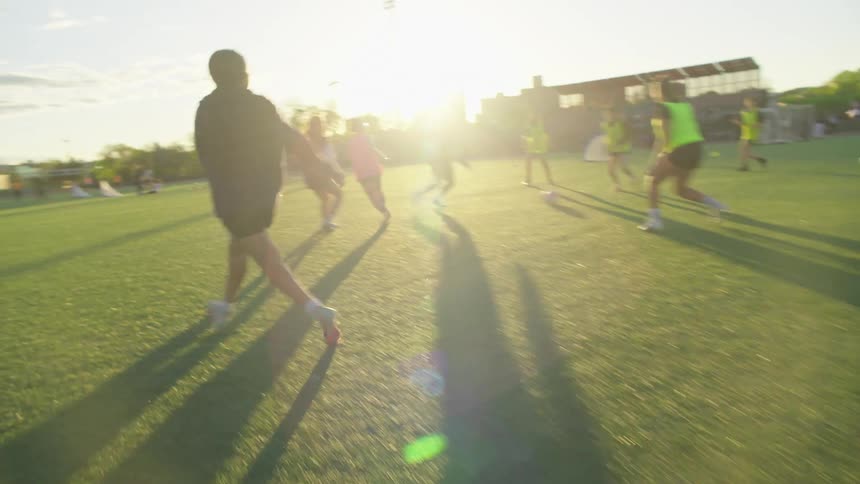 A group of young women play a friendly but competitive game of soccer in a city park at sunset, with dynamic slow-motion shots following the ball