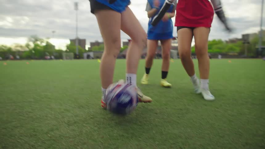A dynamic low-angle view follows a soccer ball as a team of female athletes practices passing drills on a green turf field