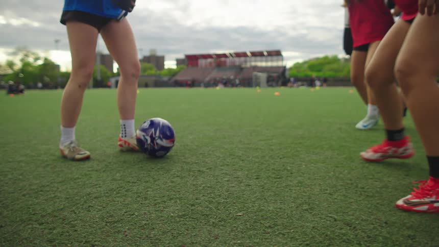 Low-angle slow motion shot of a women's soccer team practicing footwork drills with a ball on a green turf field