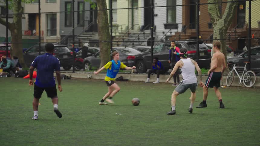 A diverse group of friends plays a casual game of soccer in slow motion on an urban park's turf field