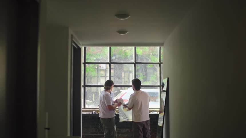 Two young men stand silhouetted in a hallway, unpacking and admiring a new graphic t-shirt in front of a large window