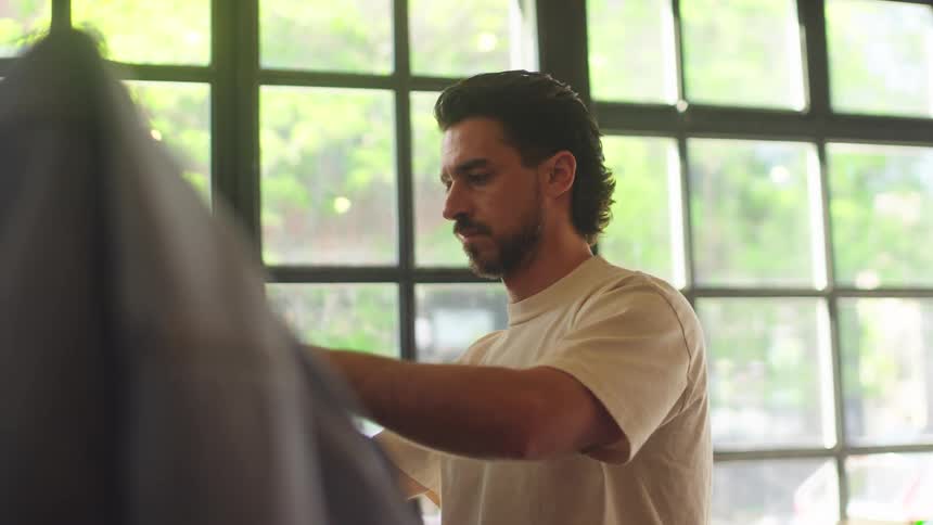 A man with a mullet hairstyle browses a rack of vintage sports jerseys in a sunlit clothing store
