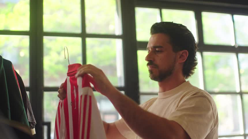 A man with a mullet thoughtfully inspects a vintage red and white striped soccer jersey in a brightly lit thrift store