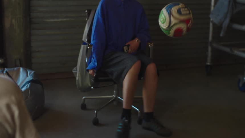 A woman in a blue shirt casually shows off her impressive soccer juggling skills while sitting in a chair in an industrial space