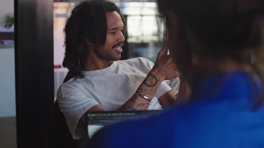 A young tattooed man with dreadlocks films his friend with a smartphone while having a conversation in a modern office