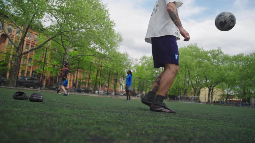 A man skillfully juggles a soccer ball with his feet in slow motion on a green turf field in a city park