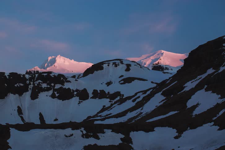 A majestic time-lapse captures the sunset casting a beautiful pink alpenglow over snowy mountain peaks as day turns to night