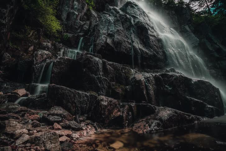 A dramatic low-angle timelapse tilts down a powerful waterfall cascading over a dark, rocky cliff in a dense forest
