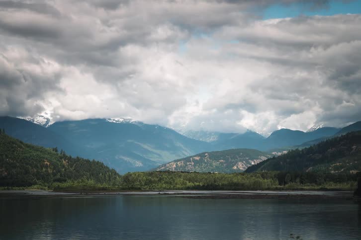 A serene landscape view of a calm lake and snow-capped mountains under a dynamic cloudy sky