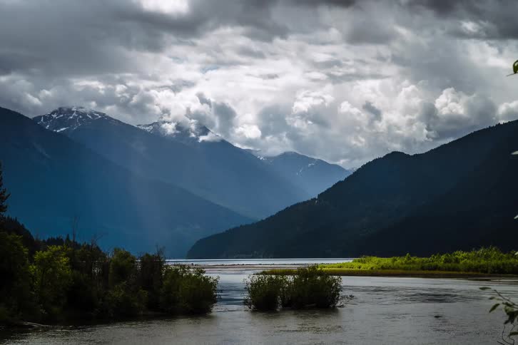 A dramatic timelapse shows sun rays breaking through stormy clouds over a majestic mountain river valley