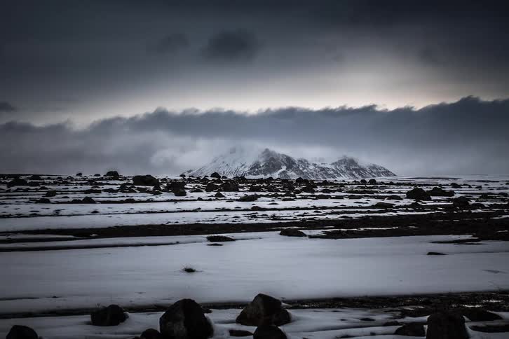 A dramatic timelapse of dark, ominous clouds moving over a vast, snowy, and rocky landscape with a distant mountain