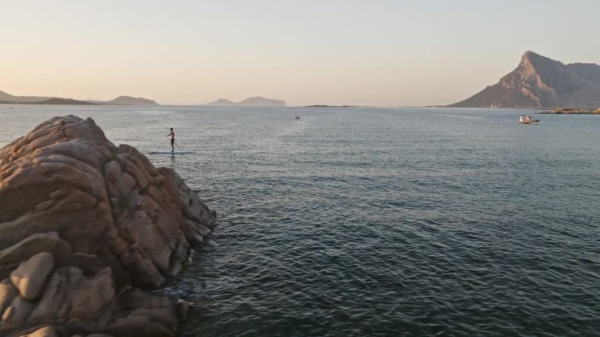 An aerial shot pans from a dramatic coastal rock formation to reveal a person on a stand-up paddleboard on the calm sea at sunrise