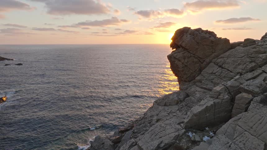 A drone pulls back along a rugged coastline as the sun rises over the ocean, creating a beautiful lens flare behind a large rock