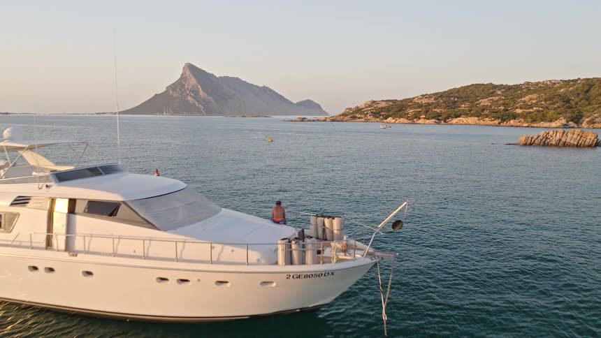 Aerial shot glides over a luxury yacht in a calm Mediterranean bay, revealing a paddleboarder and a stunning island backdrop at sunrise