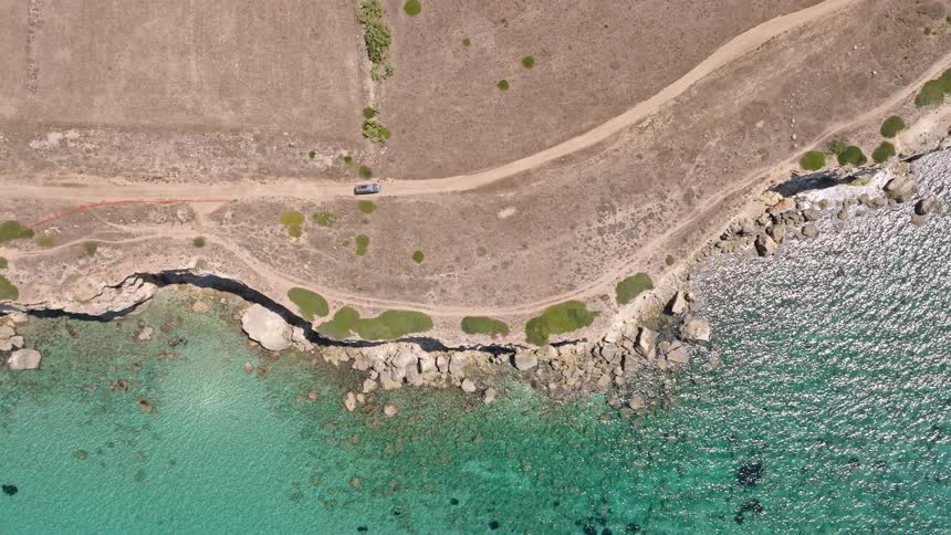 Static top-down aerial view of a car parked on a dirt road beside the stunning turquoise waters of a rocky coastline